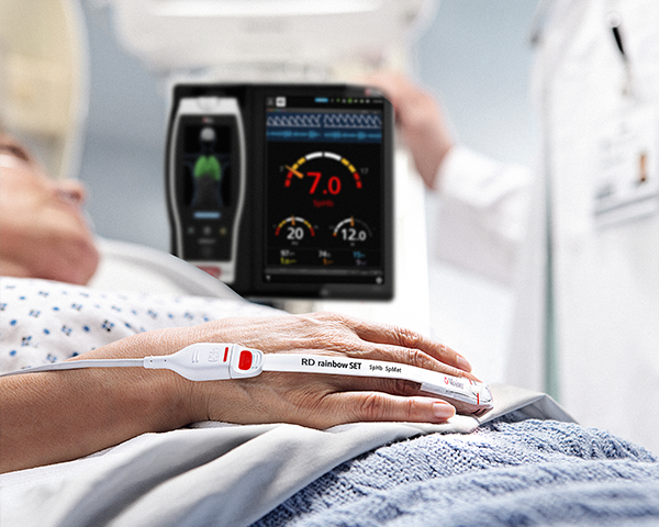 Patient sur un lit d’hôpital surveillé par un capteur Masimo avec le dispositif Root et un clinicien à l’arrière-plan, qui surveille le dispositif Root.