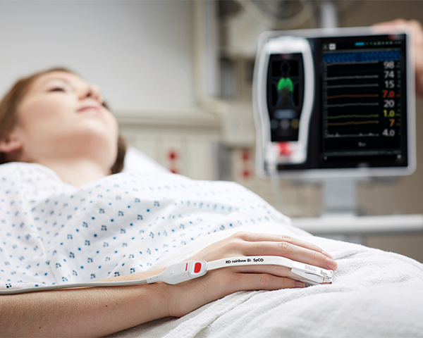 Patient endormi sur un lit d’hôpital portant un capteur de bout de doigt Masimo avec le dispositif Root à l’arrière-plan.